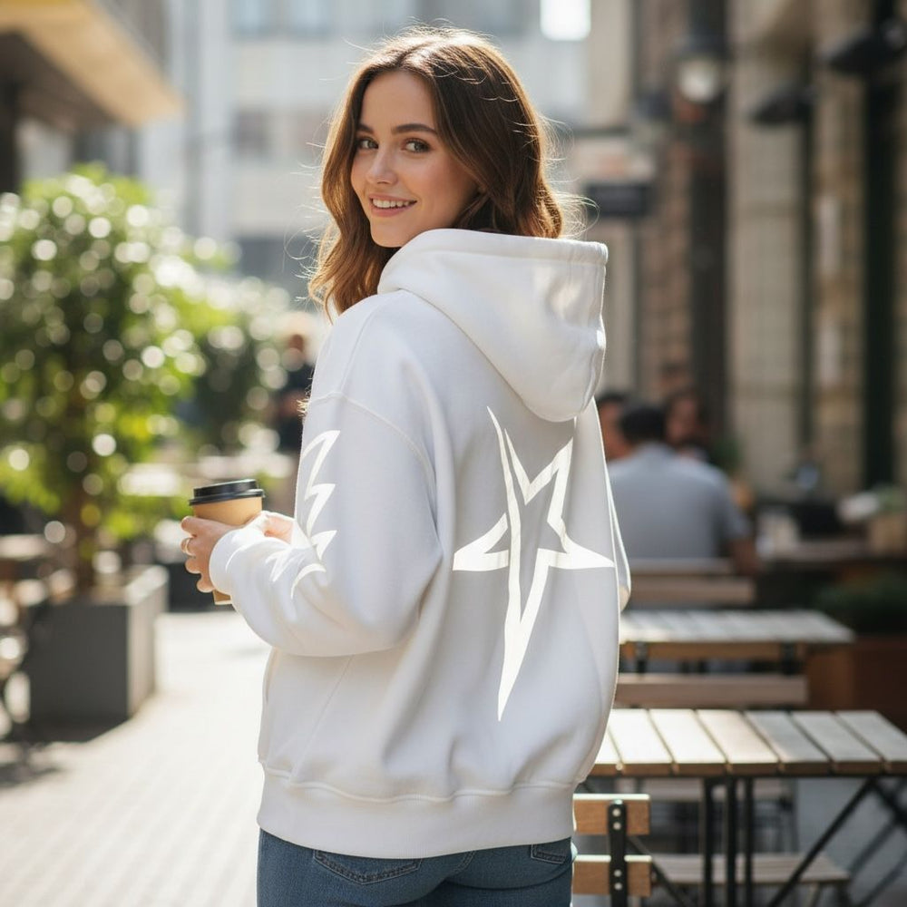 Woman wearing a white hoodie with a star design, holding a coffee cup on a city street.